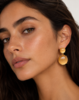 A close-up of a woman wearing a pair of chunky, double-drop dangle earrings made of warm yellow-gold-toned stone and metal, set against a clean, light off-white background.