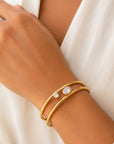 A close-up of a person's wrist wearing two thin gold bangles, one with a small clear stone, against a soft white garment background.
