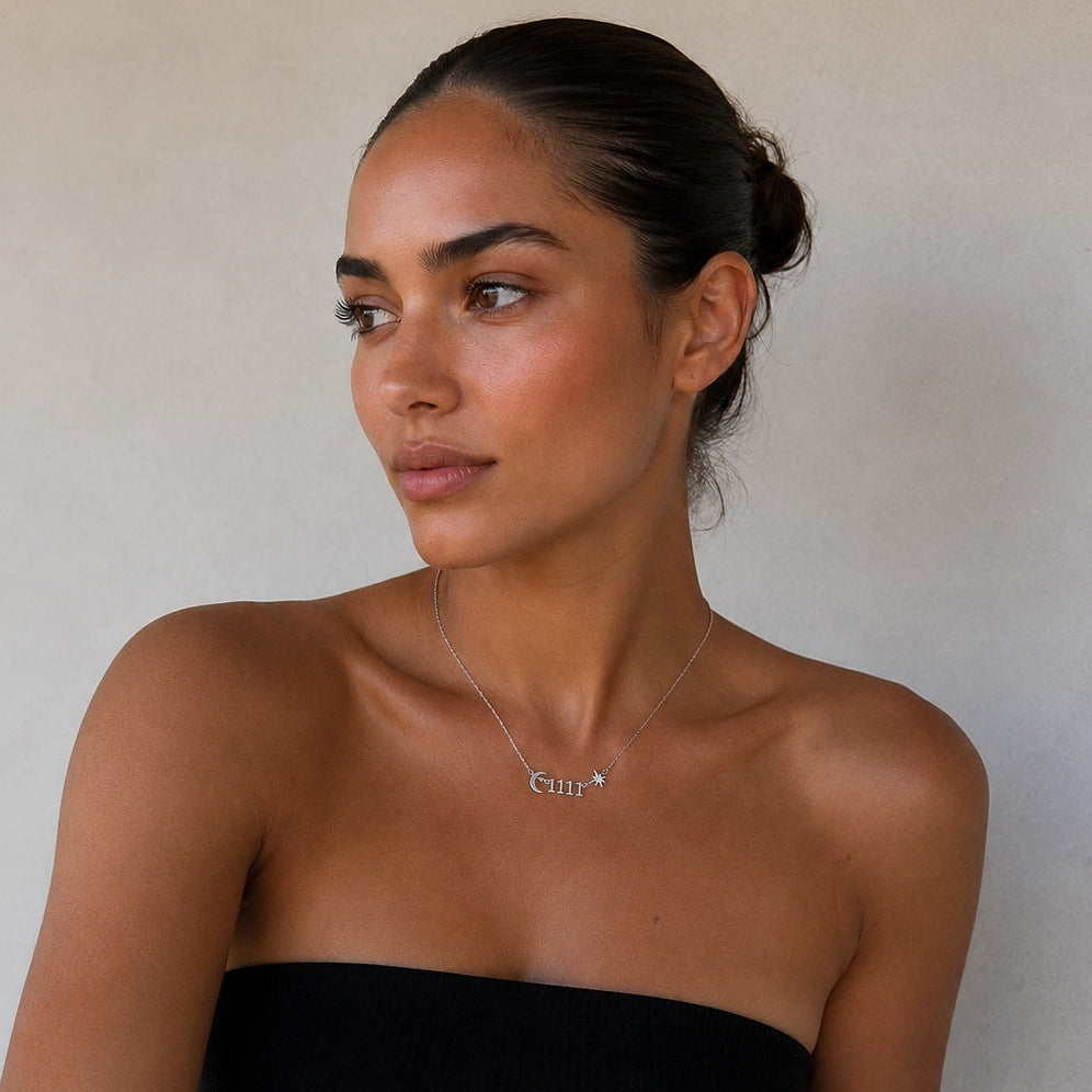 A woman wearing a black strapless tube top and white pants, wearing a delicate silver necklace with the name 'Cindy' on a light beige background.