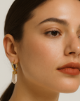 Close-up of a woman wearing gold earrings with a clear stone on a neutral background