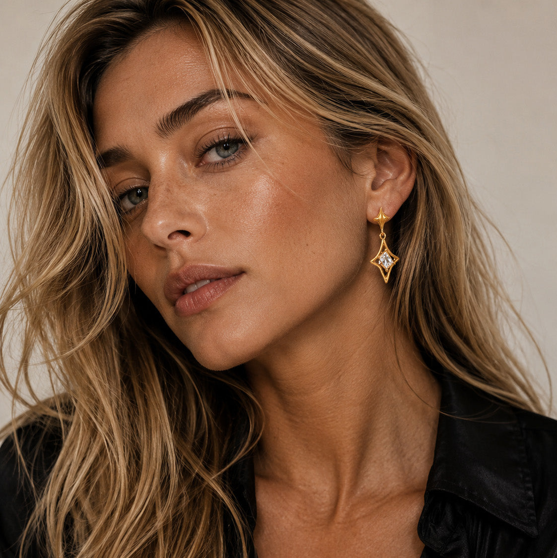 A close-up of a woman with blonde hair wearing a gold dangle earring with a clear rhinestone, paired with a black satin shirt against a neutral beige background.
