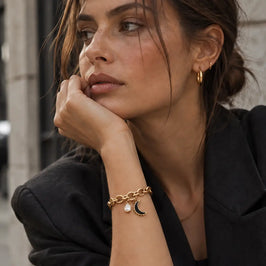 A woman wearing a gold chain bracelet with a crescent moon charm and a small pearl accent, styled with a black blazer and dark jeans.