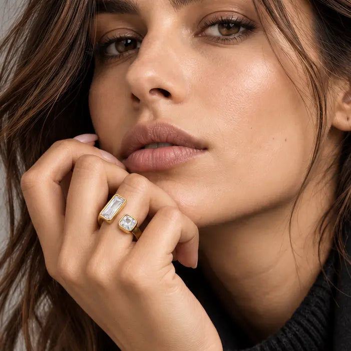 A close-up portrait of a woman wearing a silver ring with a clear gemstone on her finger, dressed in a black coat and black ribbed sweater against a neutral gray background.