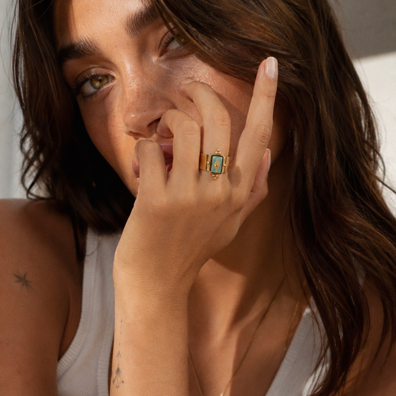 A close-up lifestyle shot of a woman wearing a gold ring with a rectangular turquoise stone on her finger, paired with a white tank top and a delicate gold chain bracelet.