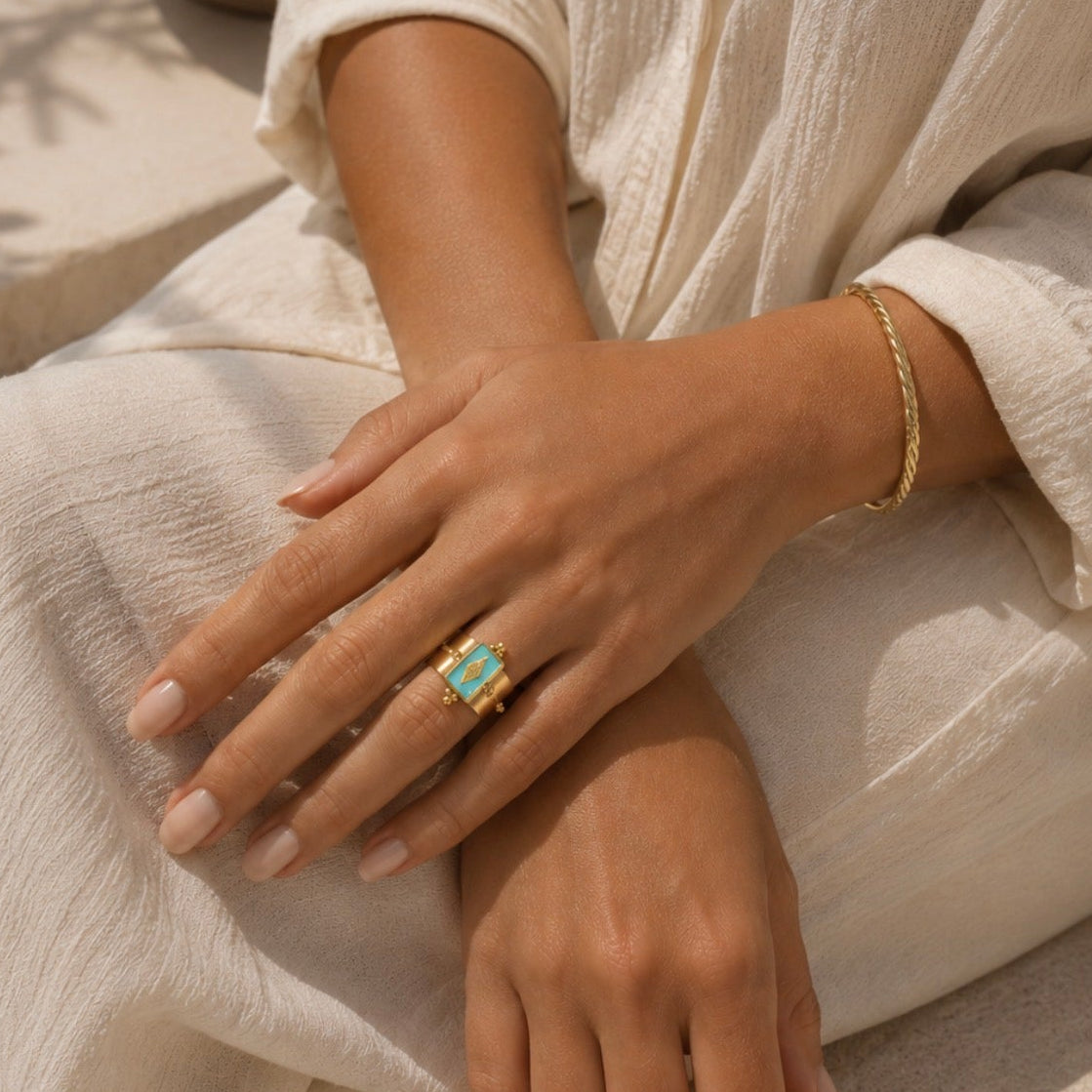 A close-up lifestyle shot of a person wearing a gold ring with a turquoise-colored rectangular stone, sitting outdoors near a pool. The person is wearing a light beige linen robe.