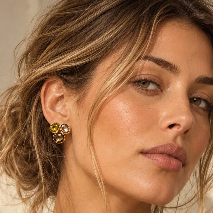 A woman with blonde-highlighted hair wearing a small gold floral stud earring, shown in a warm-toned close-up portrait against a light beige wall.