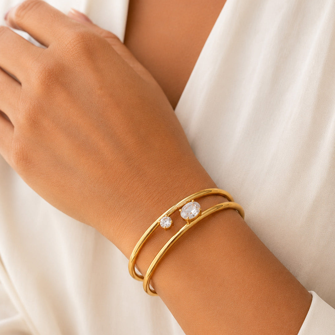A close-up of a person's wrist wearing two thin gold bangles, one with a small clear stone, against a soft white garment background.