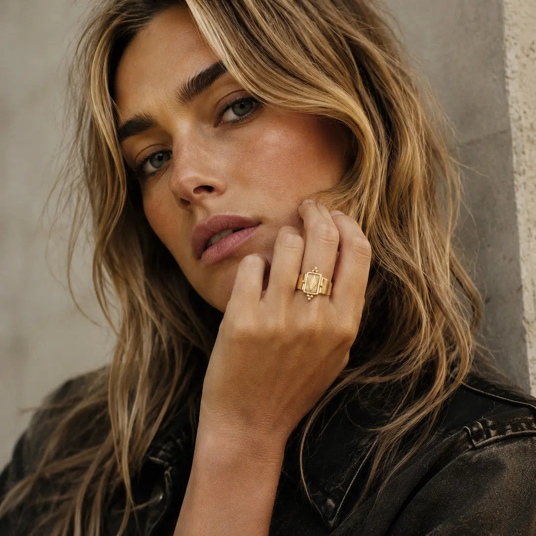 A close-up lifestyle shot of a woman with blonde-highlighted hair wearing a textured gold ring on her finger, posing in a dark denim jacket against a neutral concrete wall.