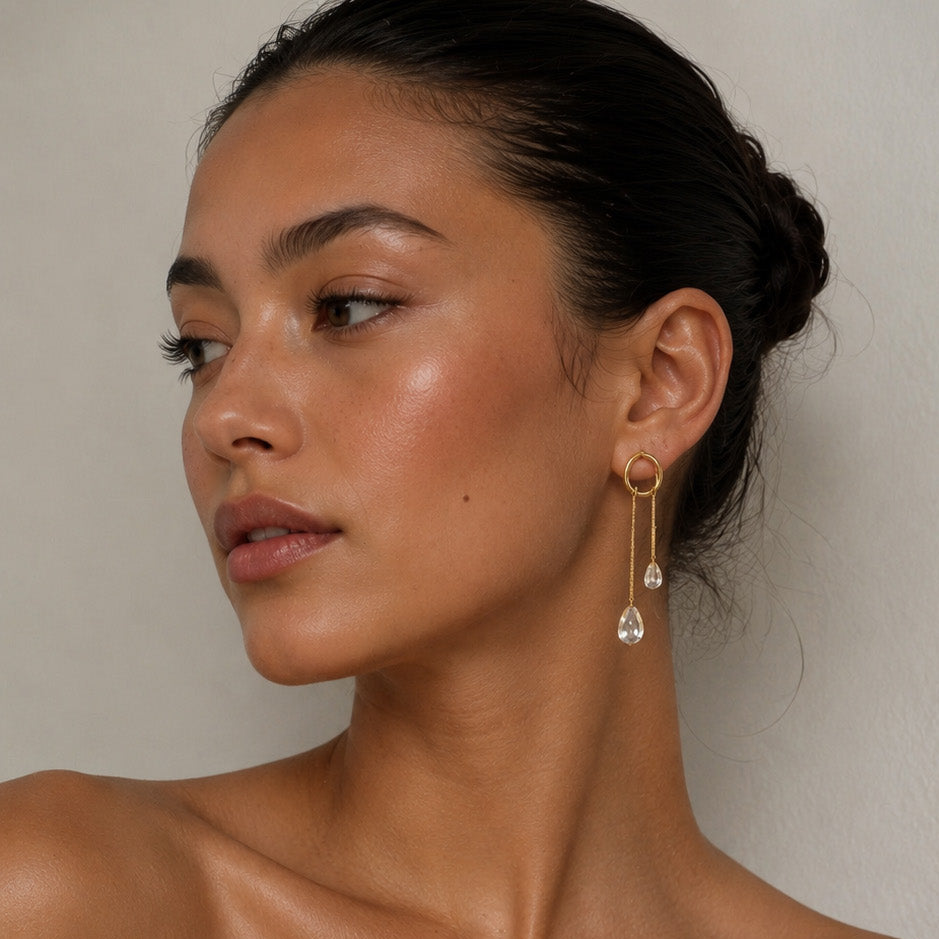 A close-up of a woman wearing a delicate gold dangle earring with a clear teardrop crystal pendant, paired with a white ribbed strapless top against a neutral light gray background.