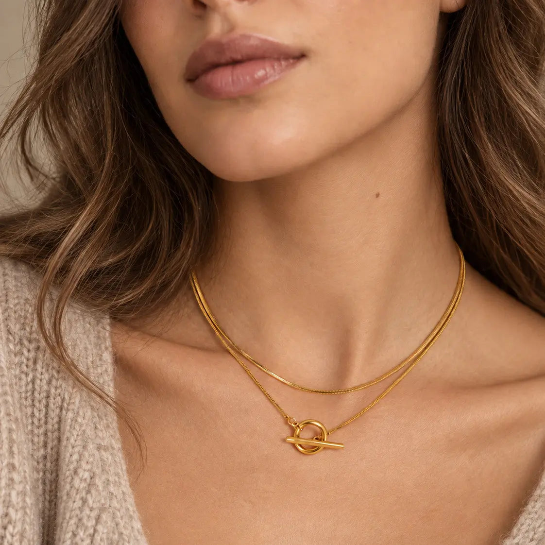 A close-up of a model wearing a delicate double-layered gold necklace with a small abstract ring pendant, styled with a beige knit sweater.
