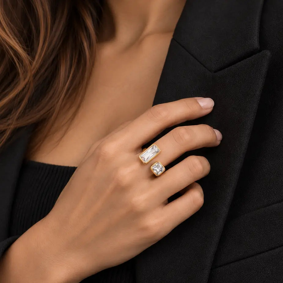 A close-up of a woman's hand wearing two gold rings with clear rectangular gemstones, styled with a black blazer and black ribbed top.