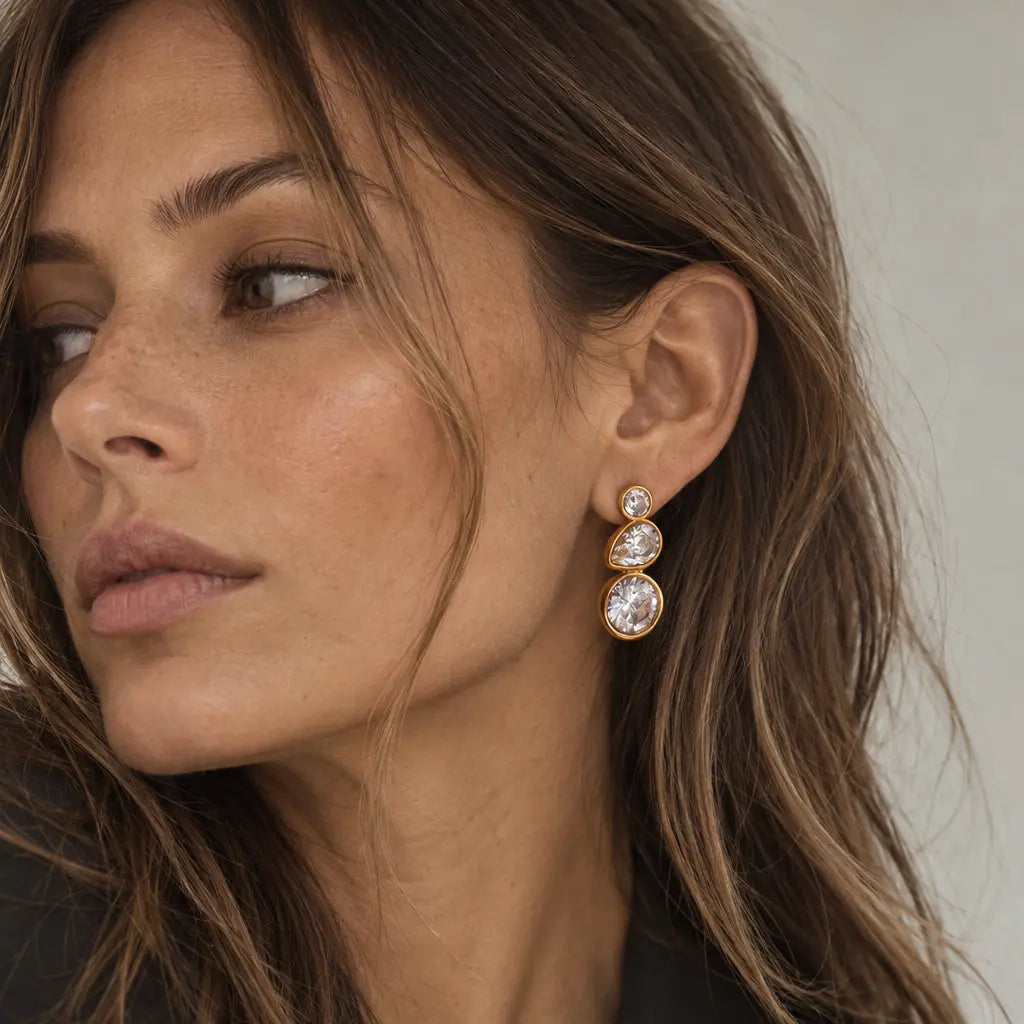 A close-up of a woman wearing a gold drop earring with three stacked round clear gemstones, paired with a black blazer.