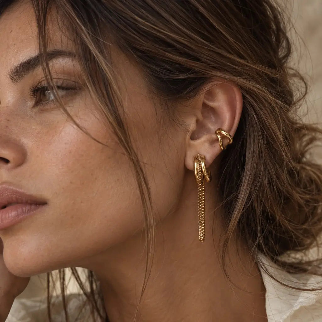 A close-up of a woman wearing a pair of gold hoop earrings with long dangling chain drops, styled with a matching gold ear cuff.