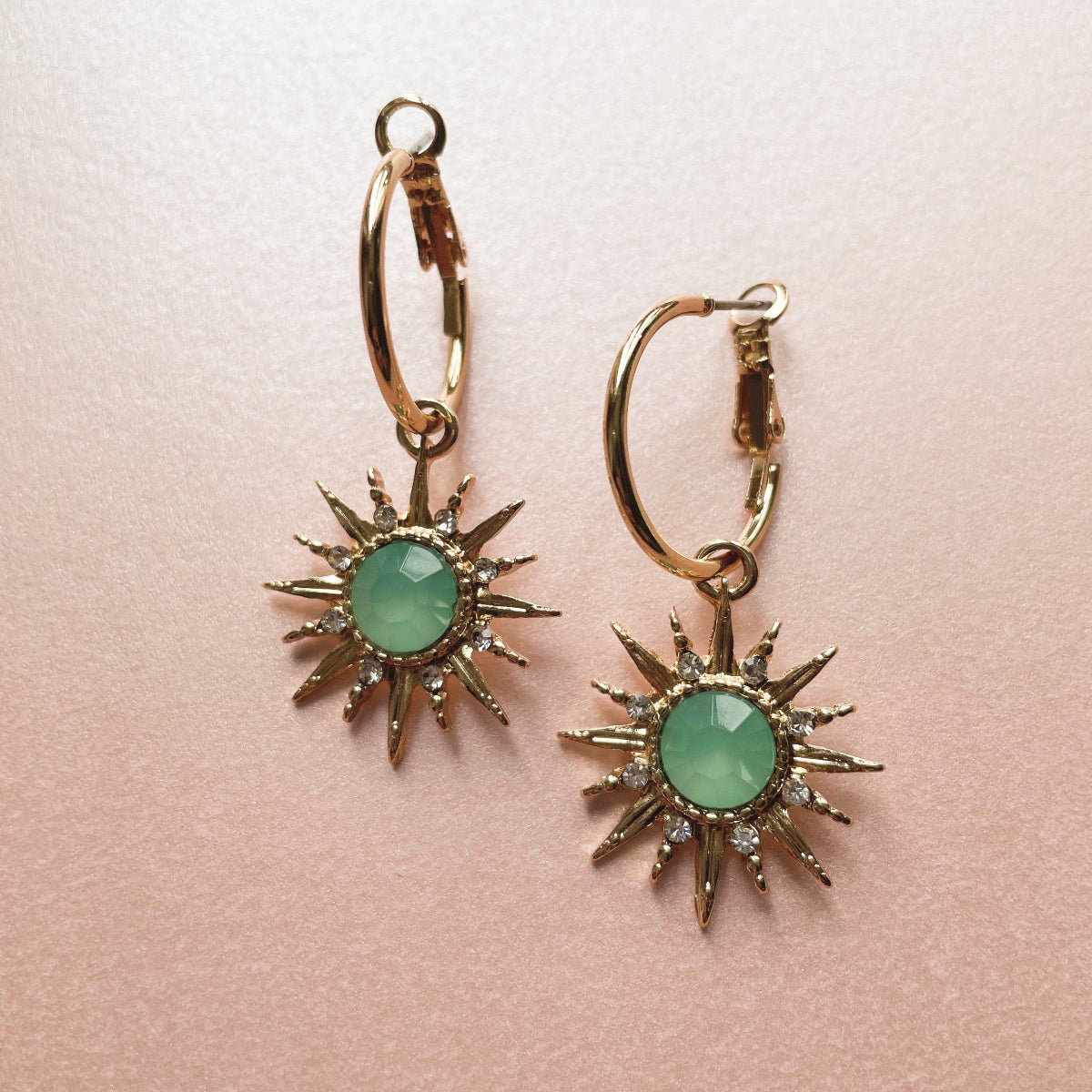 A pair of gold-tone hoop earrings with an acrylic aqua gemstone sunburst charm, displayed against a pink background.